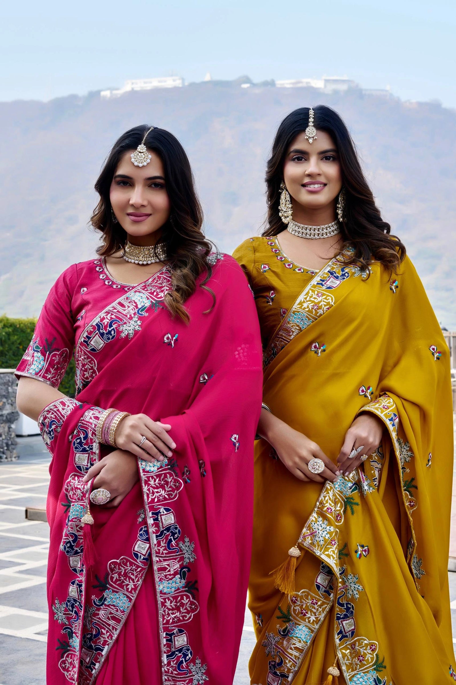 Two women in traditional sarees standing outdoors with a scenic background.