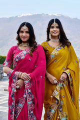 Two women in traditional sarees standing outdoors with a scenic background.
