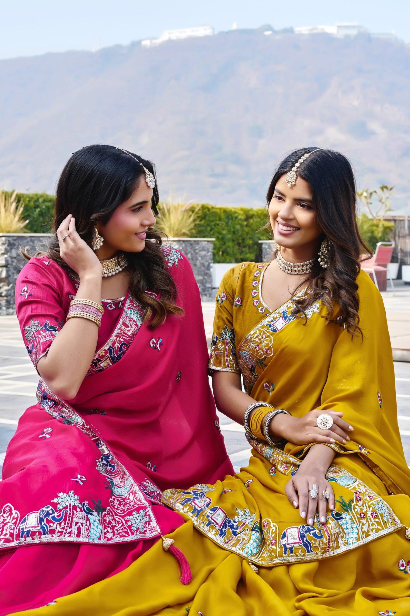 Two women in colorful traditional outfits sitting outdoors with mountains in the background