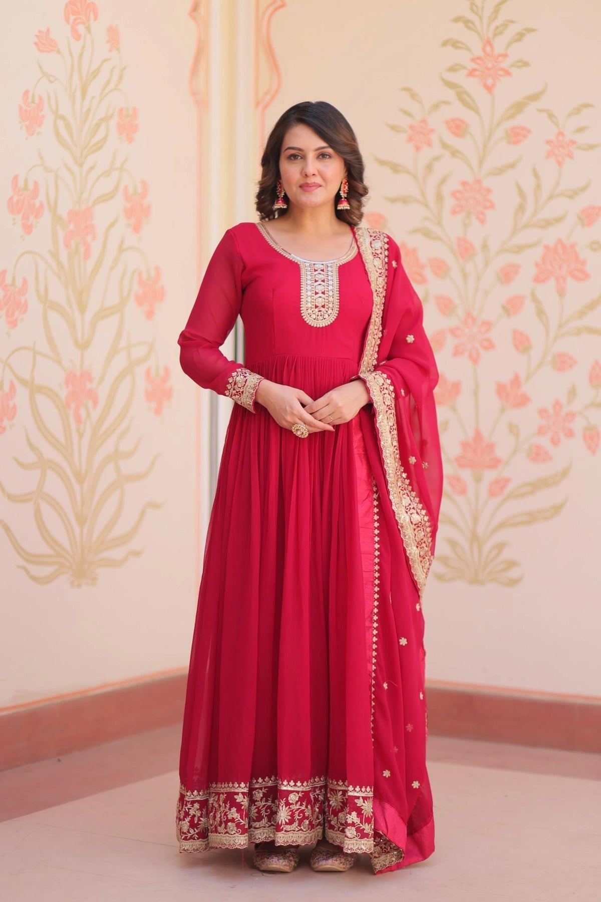 Woman wearing a red traditional outfit with gold embroidery against a floral wallpapered wall.