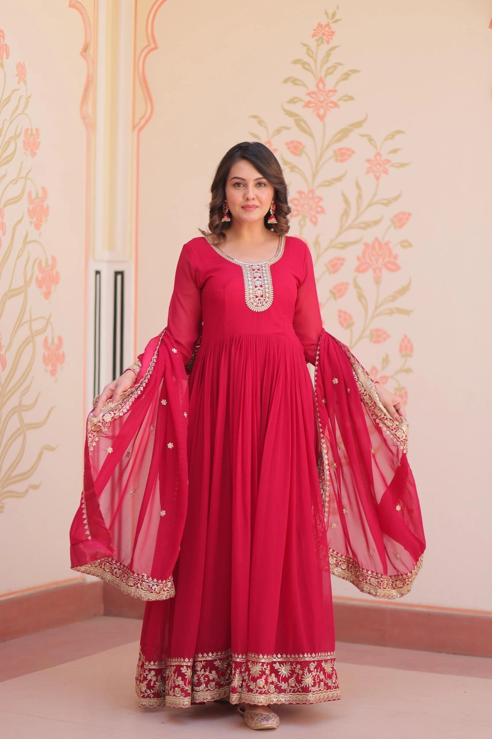 Woman wearing a red traditional outfit with floral patterns on a decorated wall.
