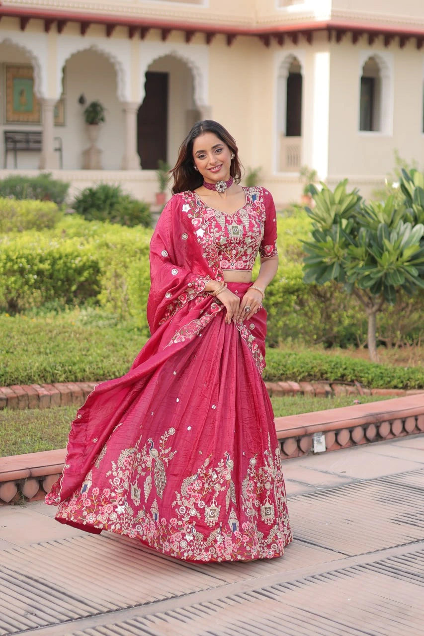 Woman in a pink embroidered saree standing outdoors with greenery and architecture in the background
