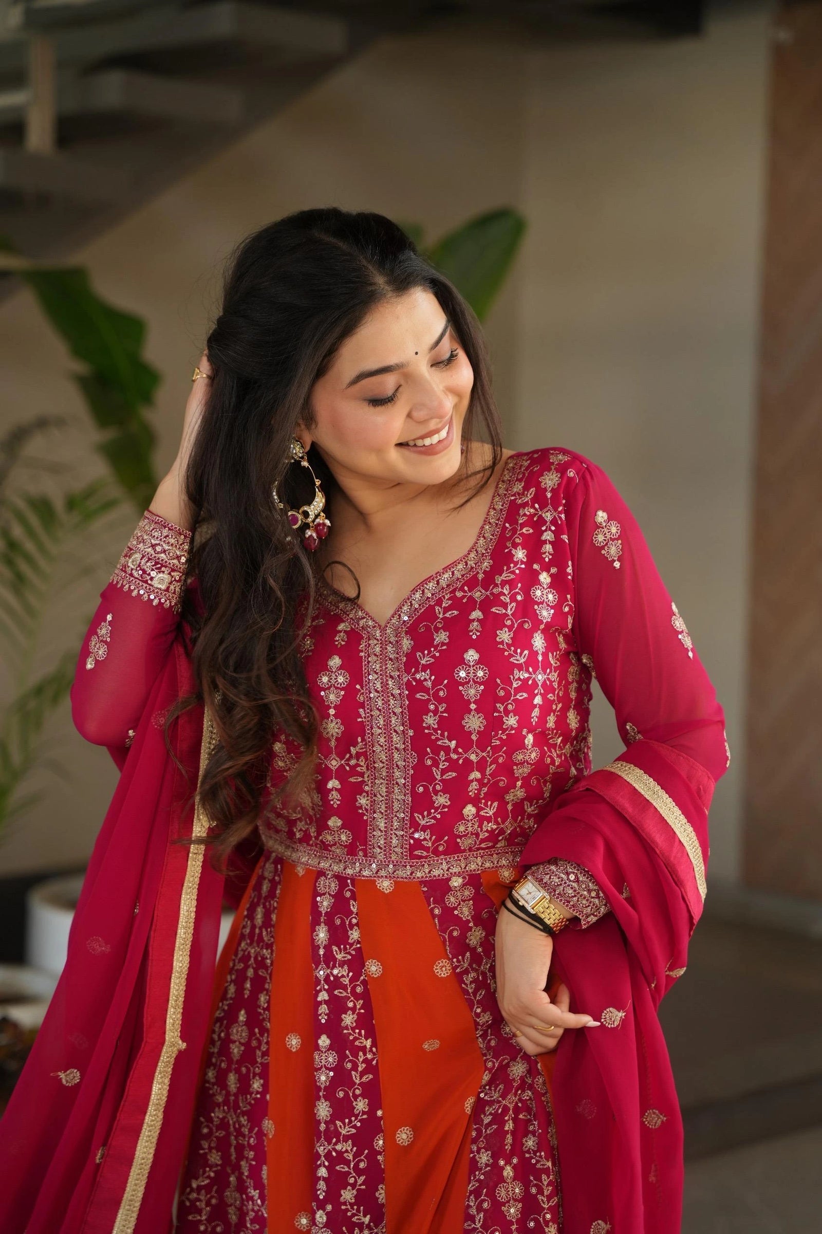 Woman in a red and orange traditional outfit with intricate patterns, standing indoors.