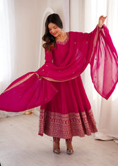 Woman in a bright pink traditional outfit with a matching dupatta, standing against a white curtain background.