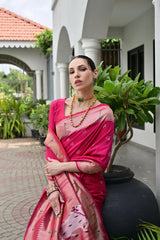 Woman in a pink saree standing outdoors with greenery and a building in the background