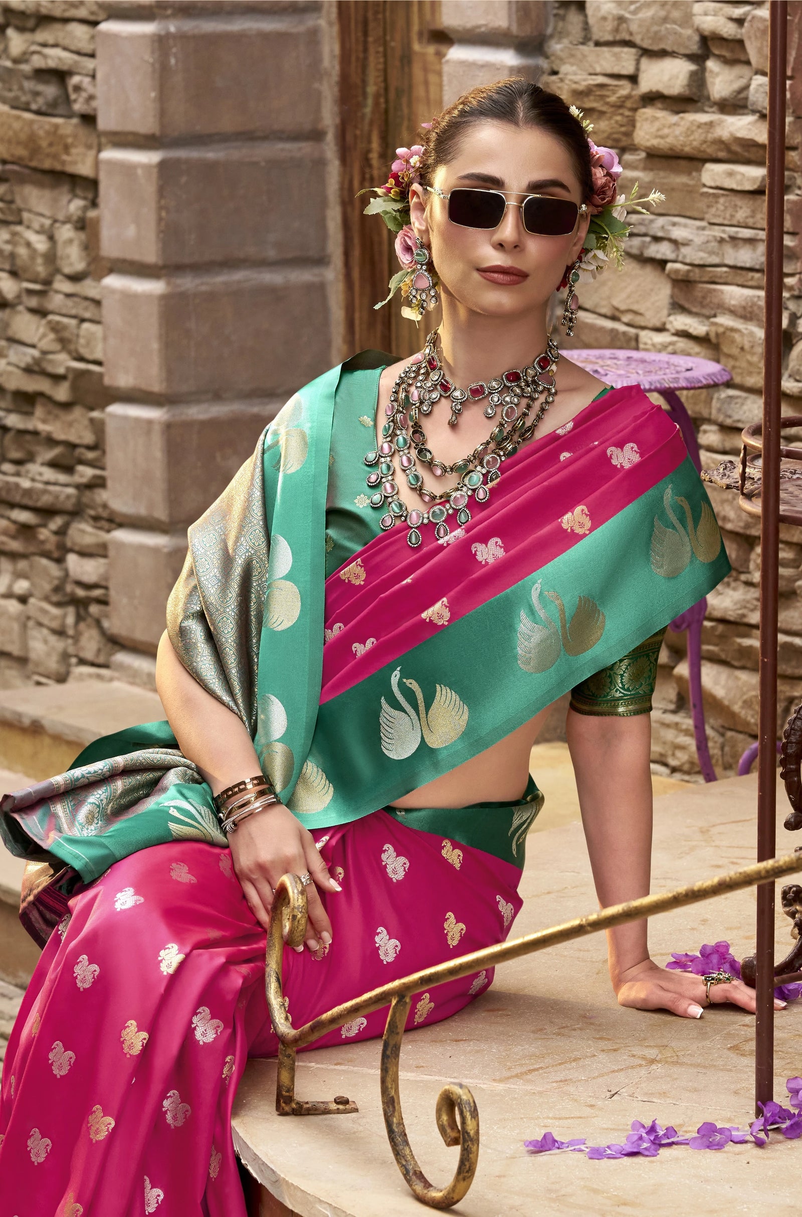 Woman in a traditional pink and green saree with jewelry, sitting outdoors.