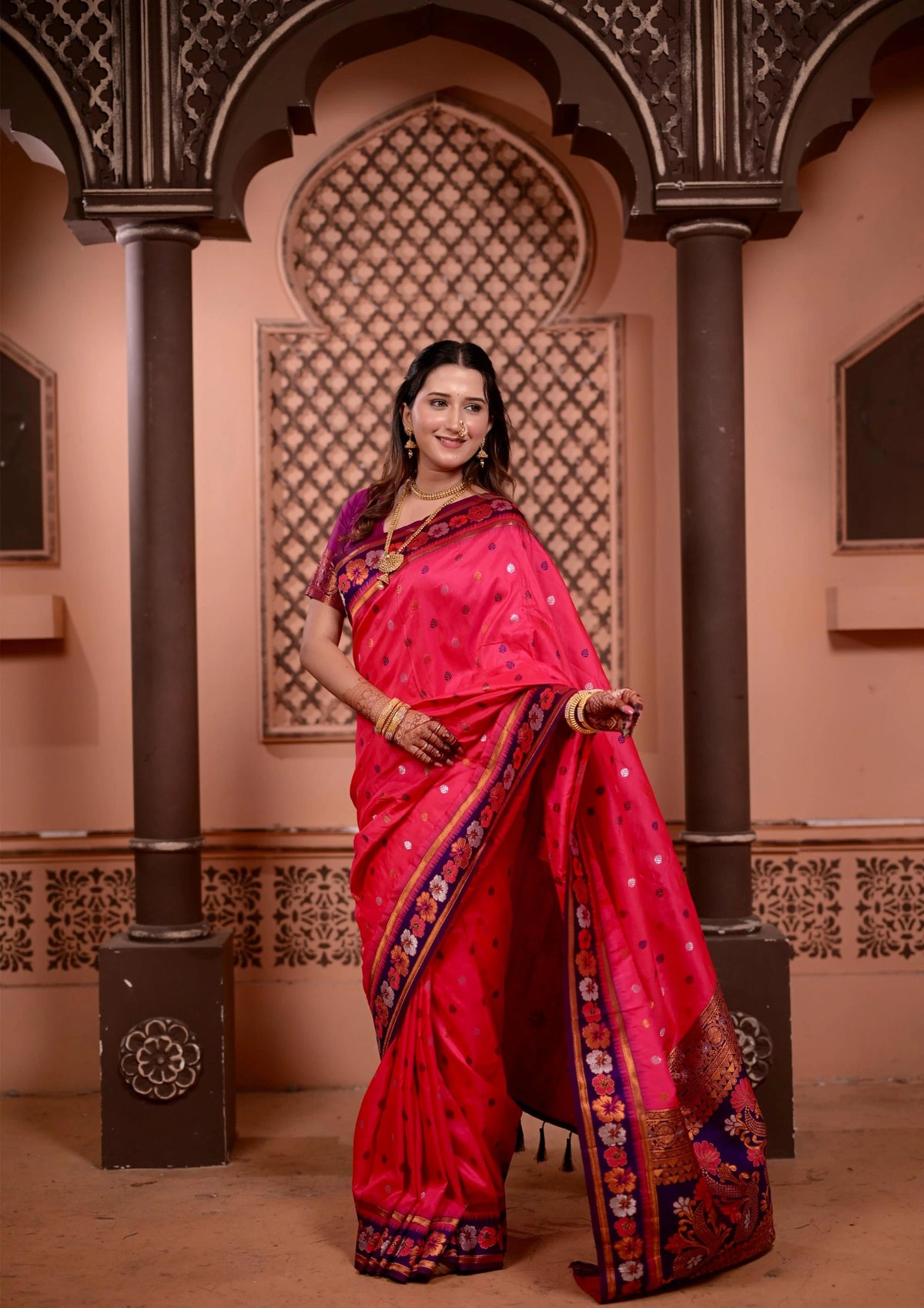 Woman in a bright pink saree with intricate patterns standing in an ornate room.