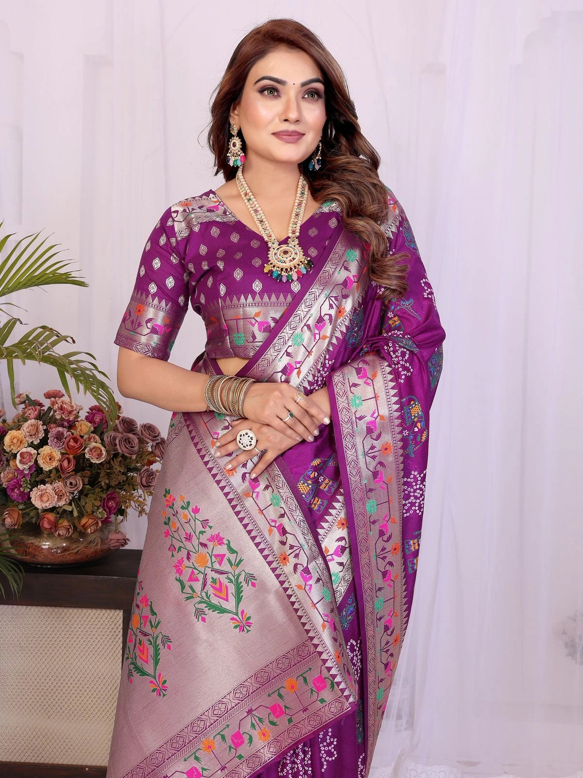 Woman wearing a purple saree with floral patterns, standing indoors with a decorative background.