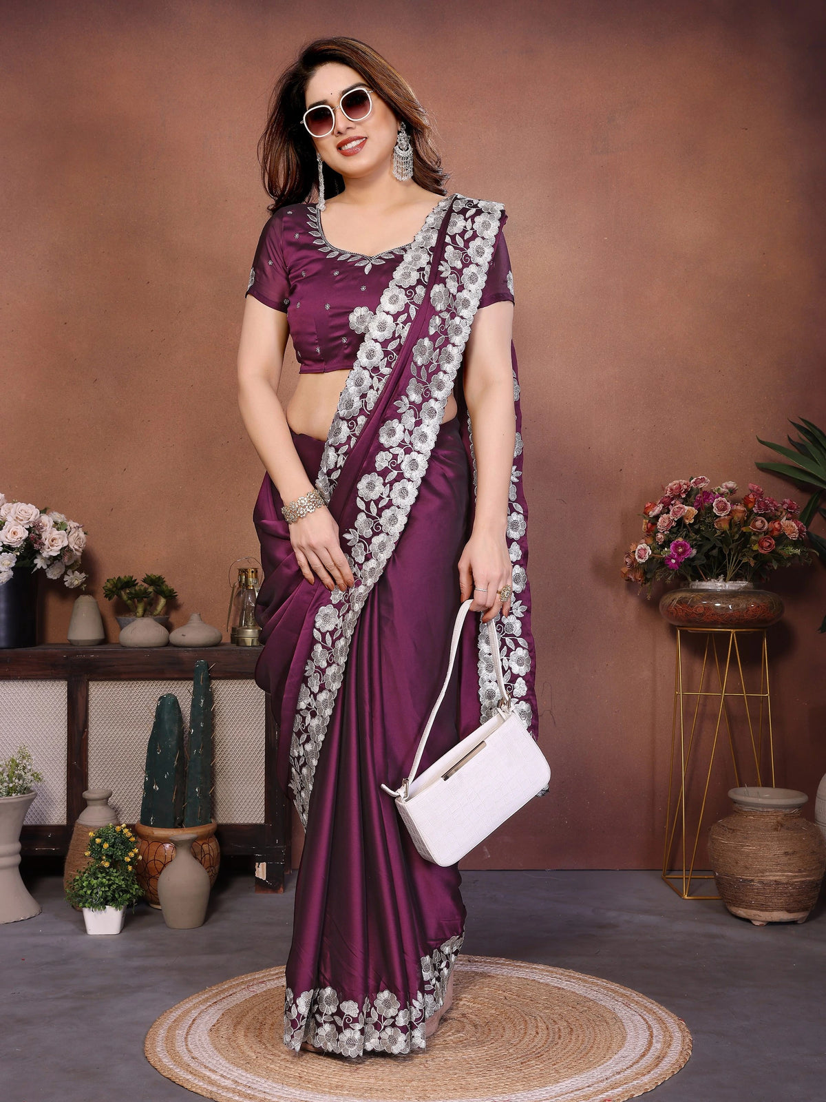 Woman in a purple saree with white floral patterns holding a white handbag, standing against a brown wall with decorative plants.
