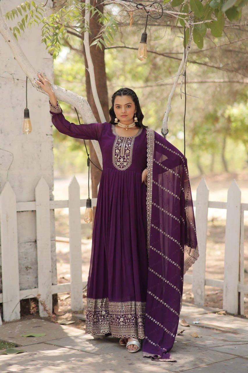 Woman in a purple traditional outfit standing outdoors with greenery and a white picket fence in the background.