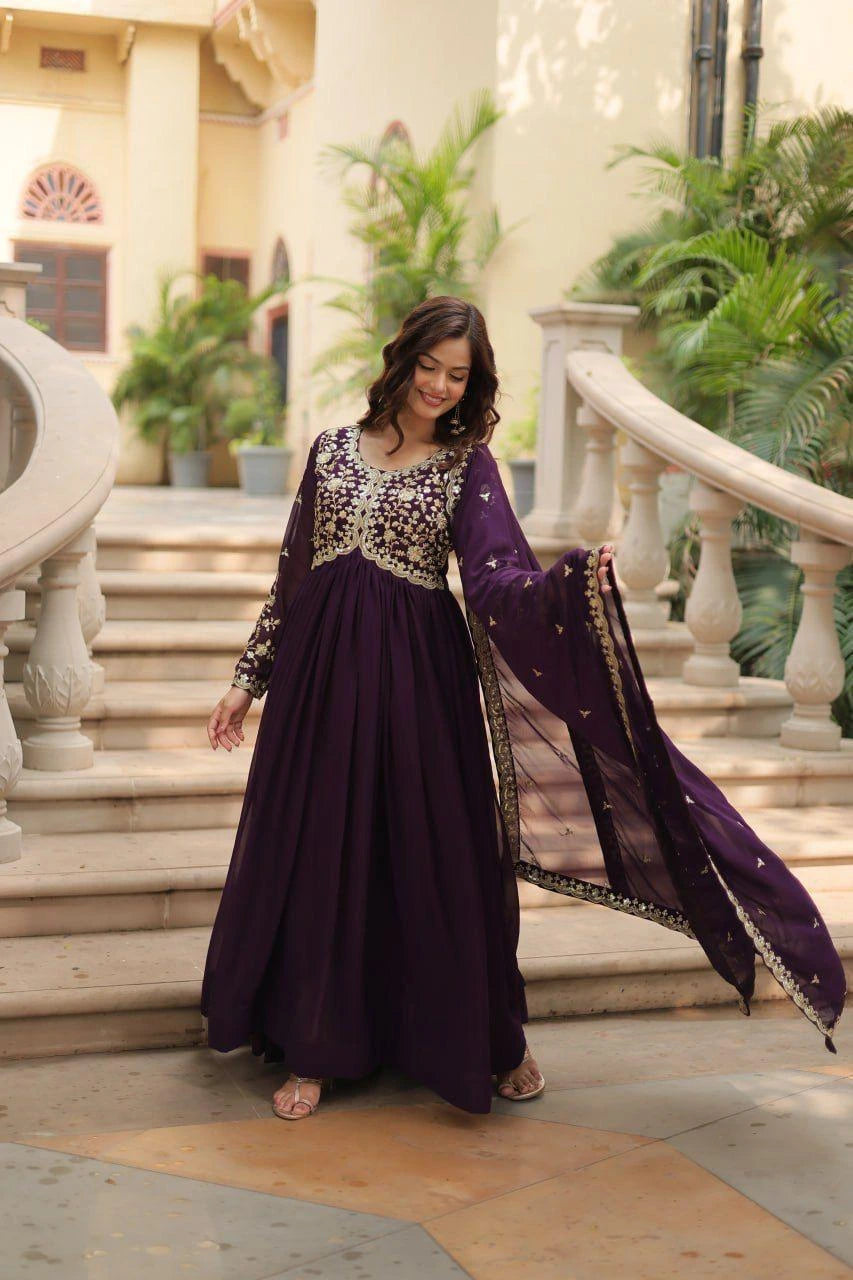 Woman in a purple and gold traditional outfit standing in an elegant indoor setting with columns and plants.