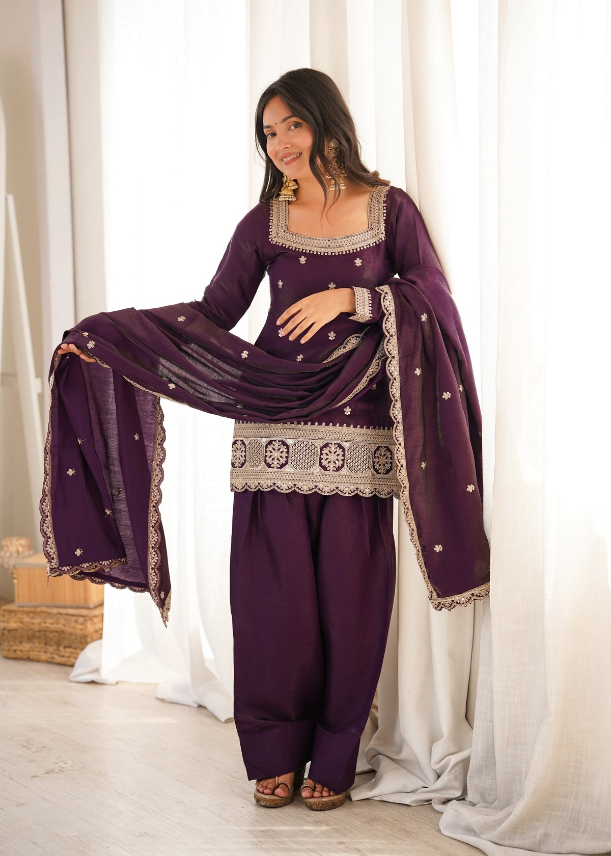 Woman in a traditional outfit with a purple dress and dupatta against a white curtain background