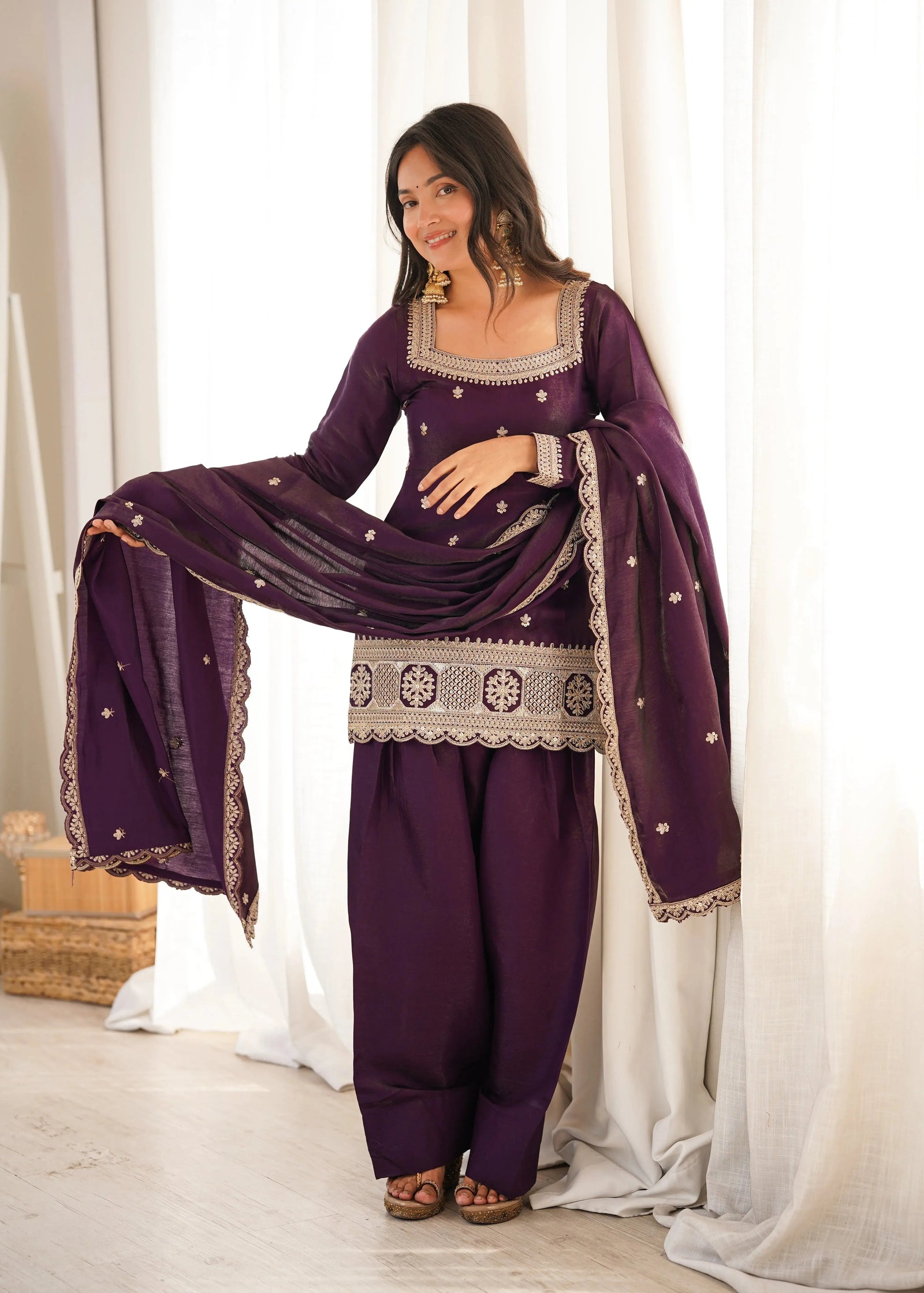 Woman in a traditional outfit with a purple dress and dupatta against a white curtain background