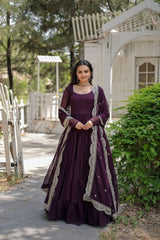 Woman in a purple traditional outfit standing outdoors with greenery and a white structure in the background.