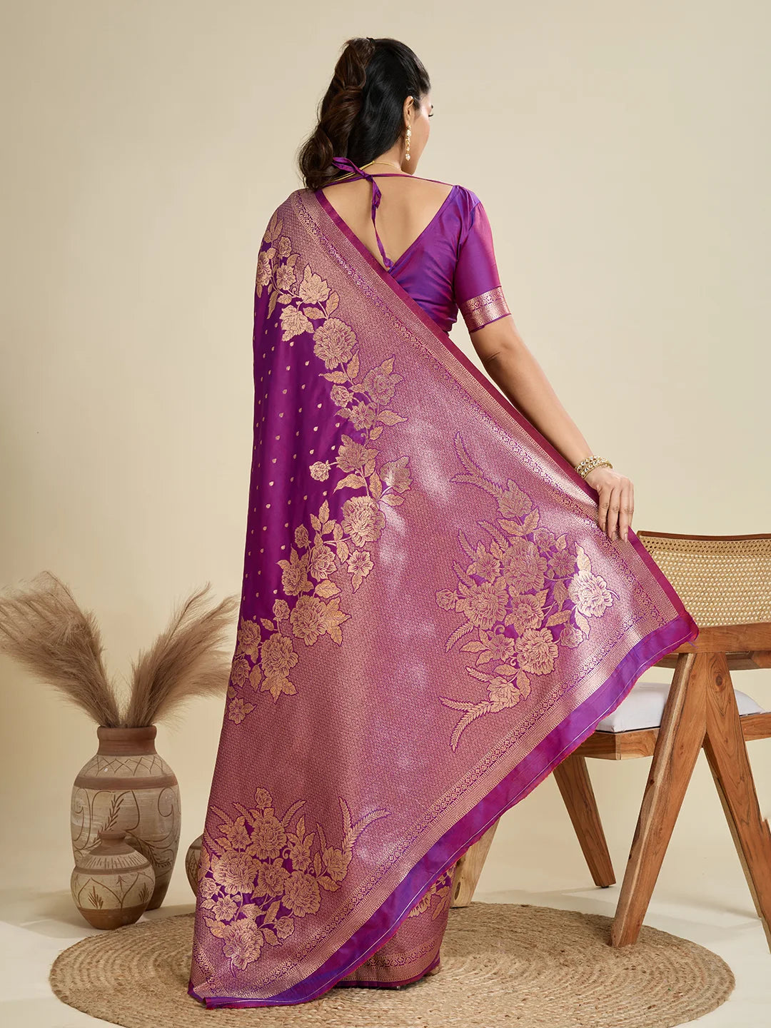 Woman wearing a pink saree with purple border, sitting on a chair against a beige background.