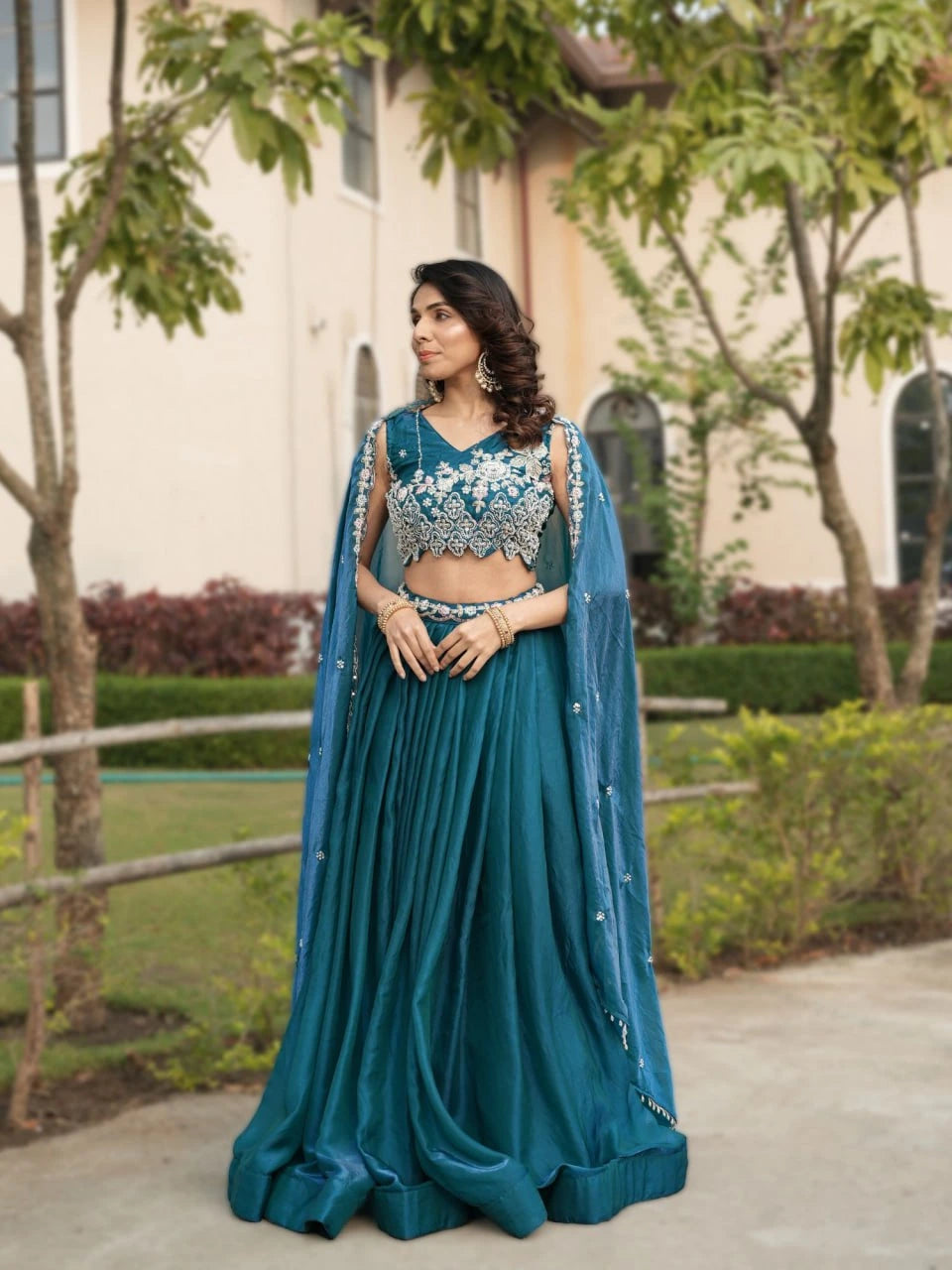 Woman in a teal traditional outfit with floral embellishments standing outdoors.
