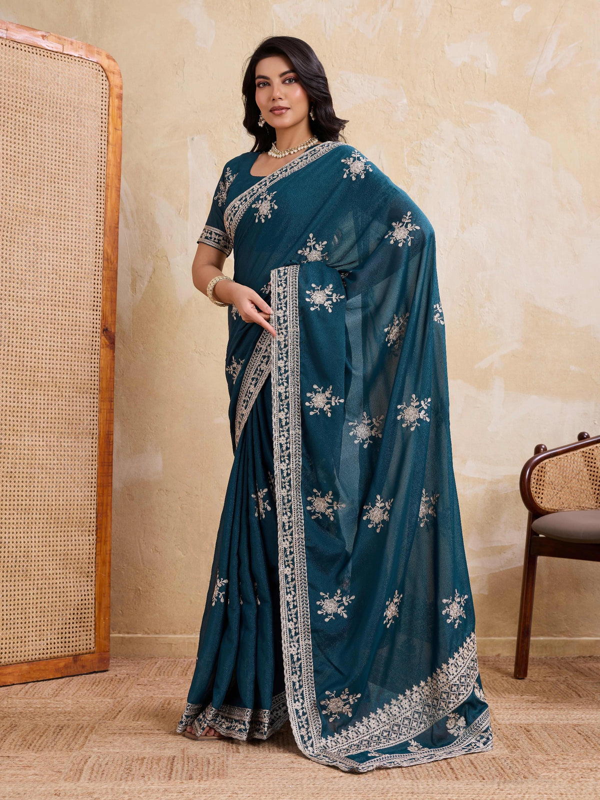 Woman wearing a teal saree with silver embroidery against a beige wall.