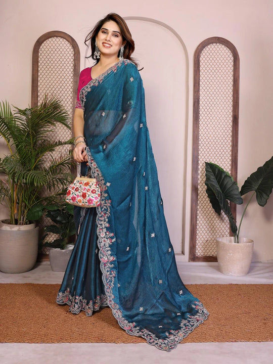 Woman wearing a blue saree with a floral design, standing indoors with plants in the background.