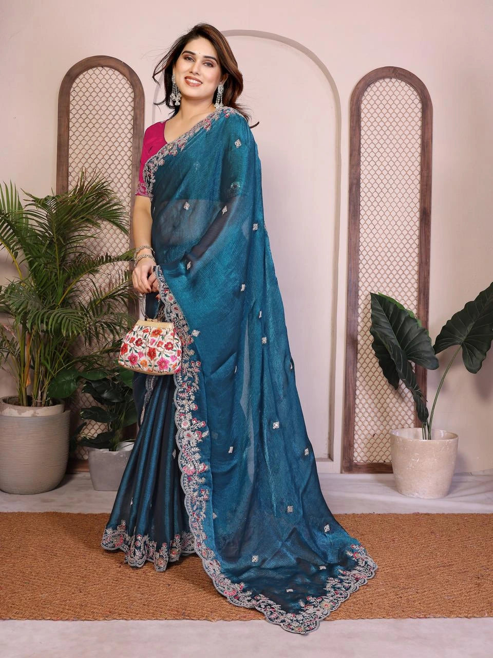 Woman wearing a blue saree with a floral design, standing indoors with plants in the background.