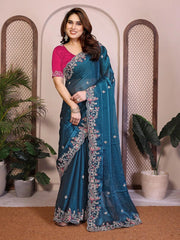 Woman wearing a blue saree with floral patterns in a decorated indoor setting.