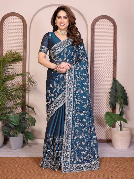 Woman wearing a blue saree with white patterns in a decorative setting with plants.