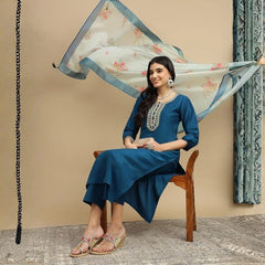 Woman in a blue traditional outfit sitting on a wooden chair with a floral dupatta draped over her head.