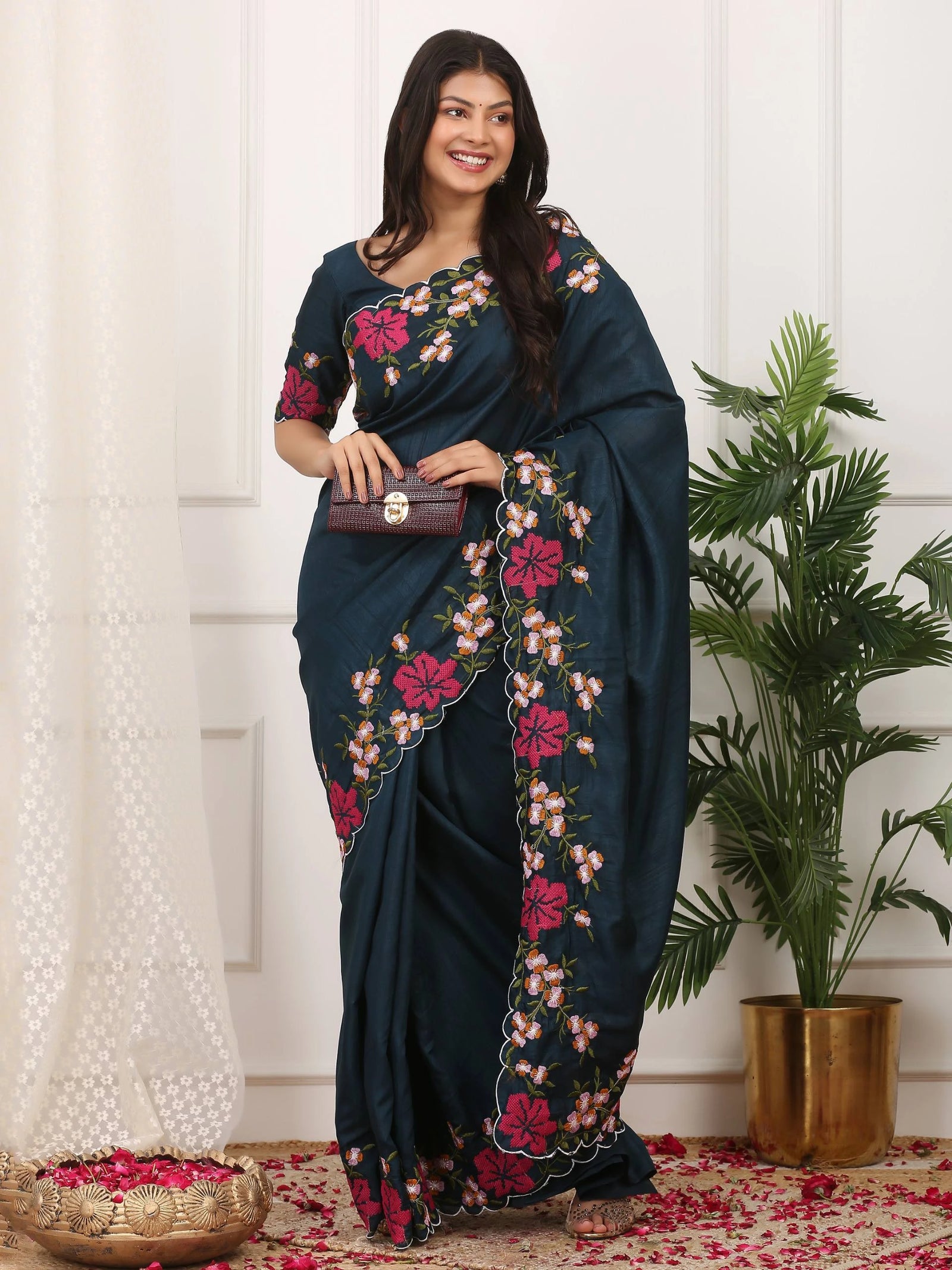 Woman wearing a dark blue saree with floral patterns in a room with white curtains and a plant.