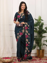 Woman wearing a dark blue saree with floral patterns in a room with white curtains and a plant.