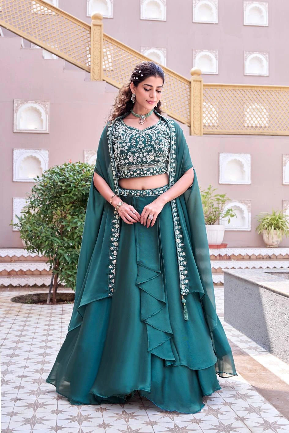 Woman in a teal traditional outfit standing in an outdoor setting with plants and architectural elements.
