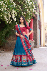Woman in a traditional blue and red saree standing outdoors with flowers and a building in the background