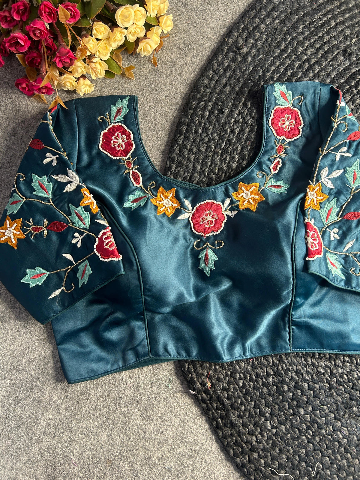 Front view of a teal blue silk-satin blouse featuring a deep U-neckline and short sleeves. The garment is decorated with vibrant hand-embroidery, including ruby red and mustard yellow flowers, light turquoise leaves, and intricate gold thread vines.