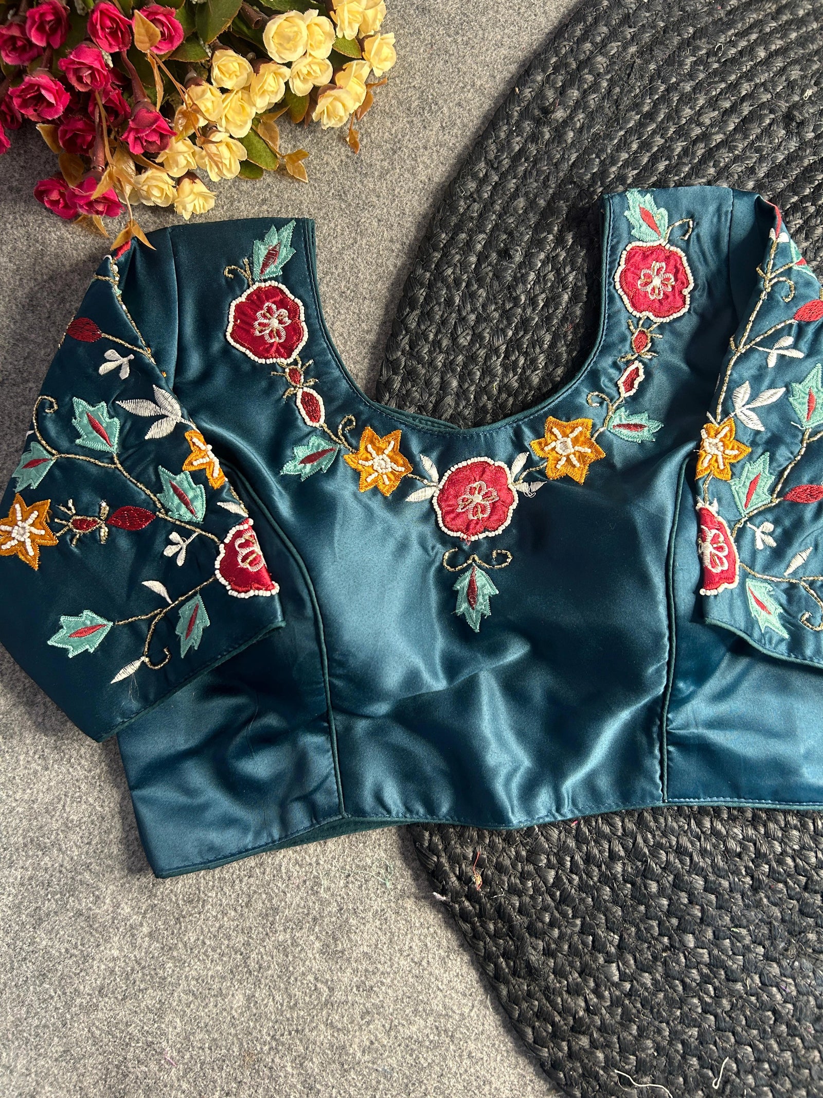 Front view of a teal blue silk-satin blouse featuring a deep U-neckline and short sleeves. The garment is decorated with vibrant hand-embroidery, including ruby red and mustard yellow flowers, light turquoise leaves, and intricate gold thread vines.