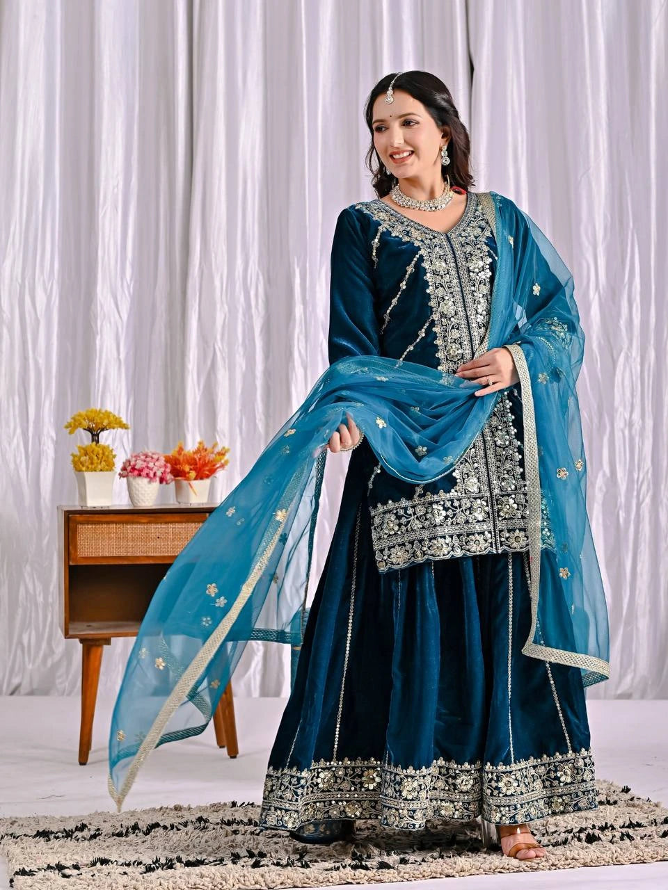 Woman in a traditional blue and silver outfit with a matching dupatta against a white curtain background.