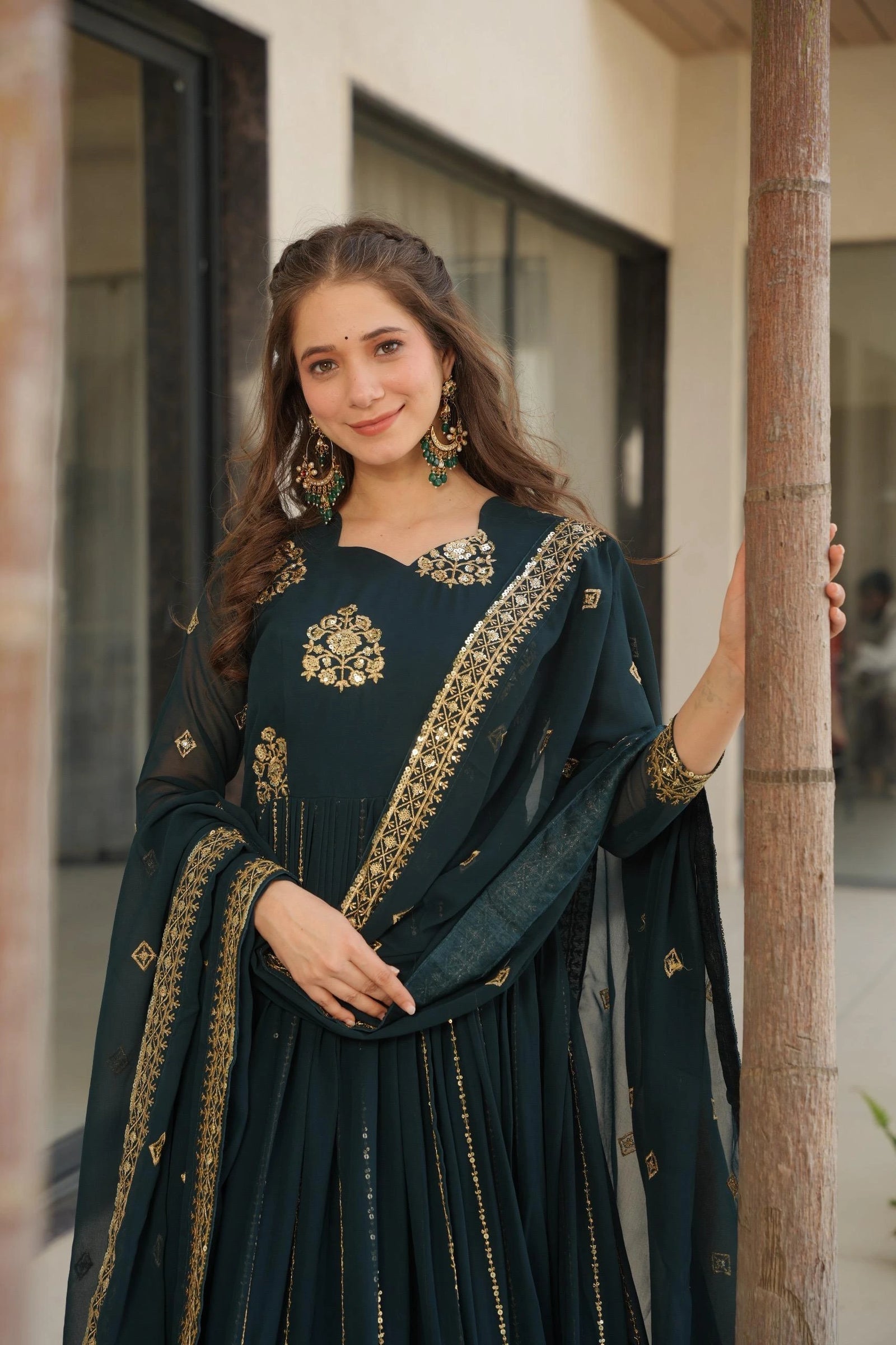 Woman in a dark green traditional outfit with gold embroidery standing against a building.