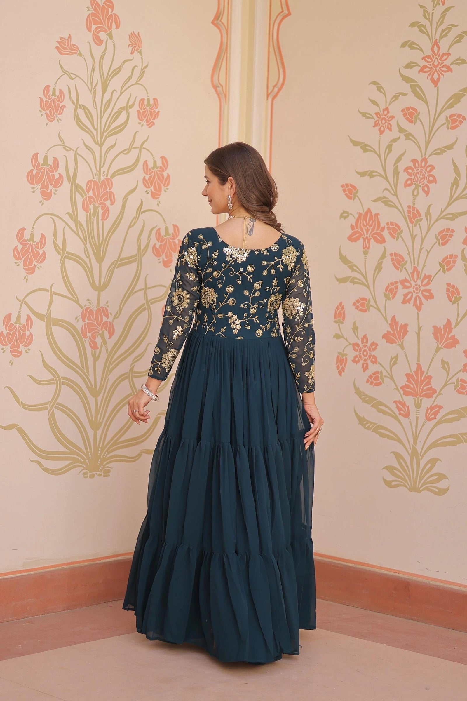 Woman in a dark blue traditional outfit with floral patterns against a decorative wall.