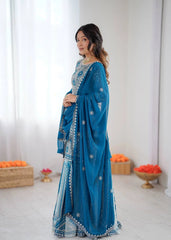 Woman wearing a blue embroidered saree in a minimalistic setting with white walls and orange flowers.