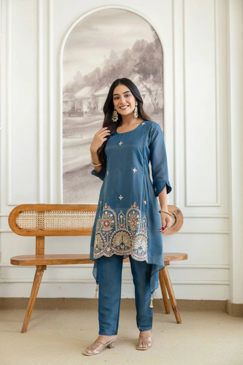 Woman in a blue traditional outfit standing in a room with a wooden bench and decorative wall.