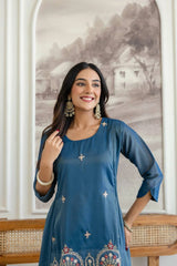 Woman wearing a blue embroidered dress standing in front of a decorative wall.