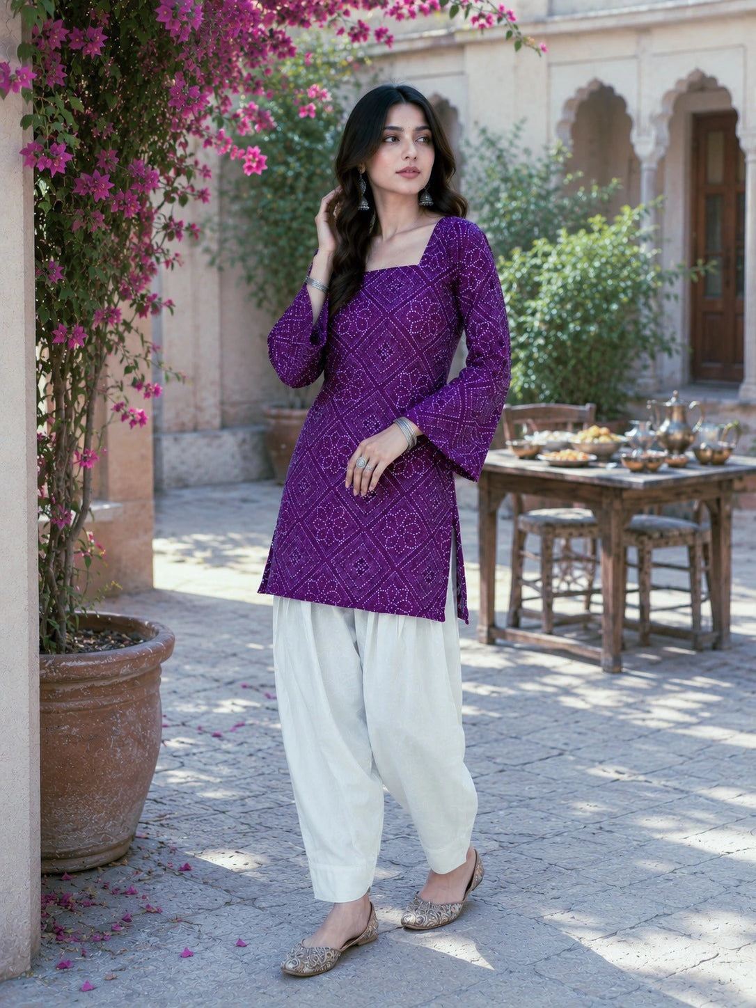 Woman in a purple top and white pants standing in an outdoor setting with flowers and a table in the background.