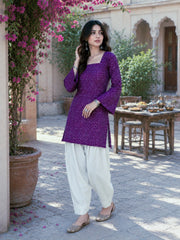 Woman in a purple top and white pants standing in an outdoor setting with flowers and a table in the background.