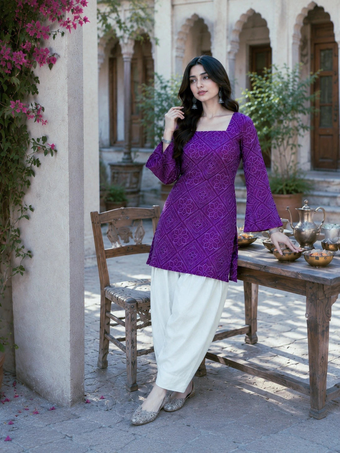 Woman in a purple top and white pants standing in an outdoor setting with a table and chairs.