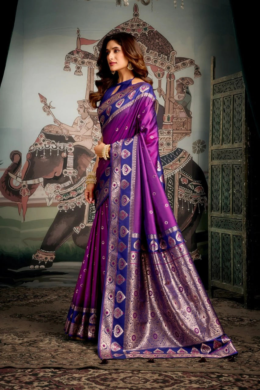 Woman in a purple and blue saree standing in front of an ornate wall with elephant and architectural elements.