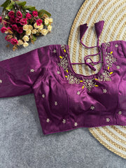 The front of the plum silk blouse showcases a curved V-neckline with the same detailed pearl and gold floral embroidery and yellow accents. The garment is tailored with structured princess-line seams and is adorned with scattered embroidered pearl motifs. The blouse is laid flat on a grey textured background next to a round jute mat and a bouquet of pink and white flowers.
