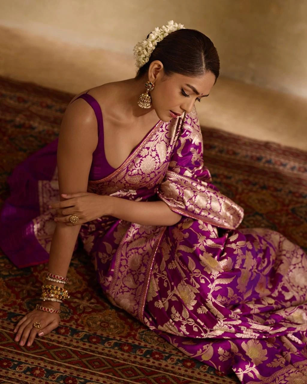 Woman in a traditional purple and gold saree sitting on a patterned rug.