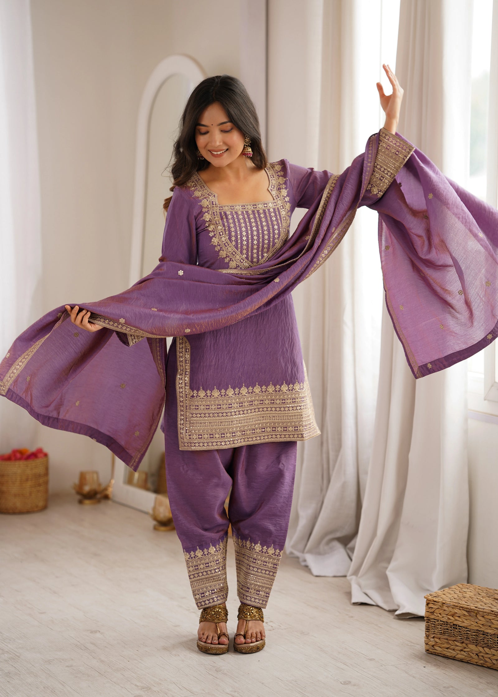 Woman in a traditional purple outfit with gold embroidery standing in a room with white curtains.
