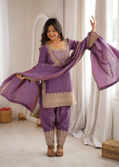 Woman in a traditional purple outfit with gold embroidery standing in a room with white curtains.