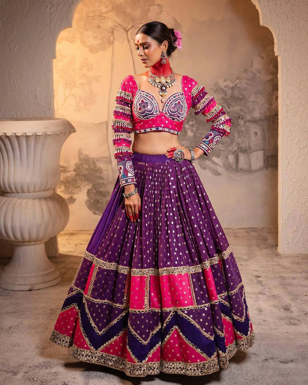Woman wearing a traditional outfit with a pink and purple top and purple skirt in an architectural setting.