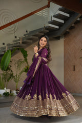 Woman in a purple and gold traditional outfit standing indoors with plants and a staircase in the background.