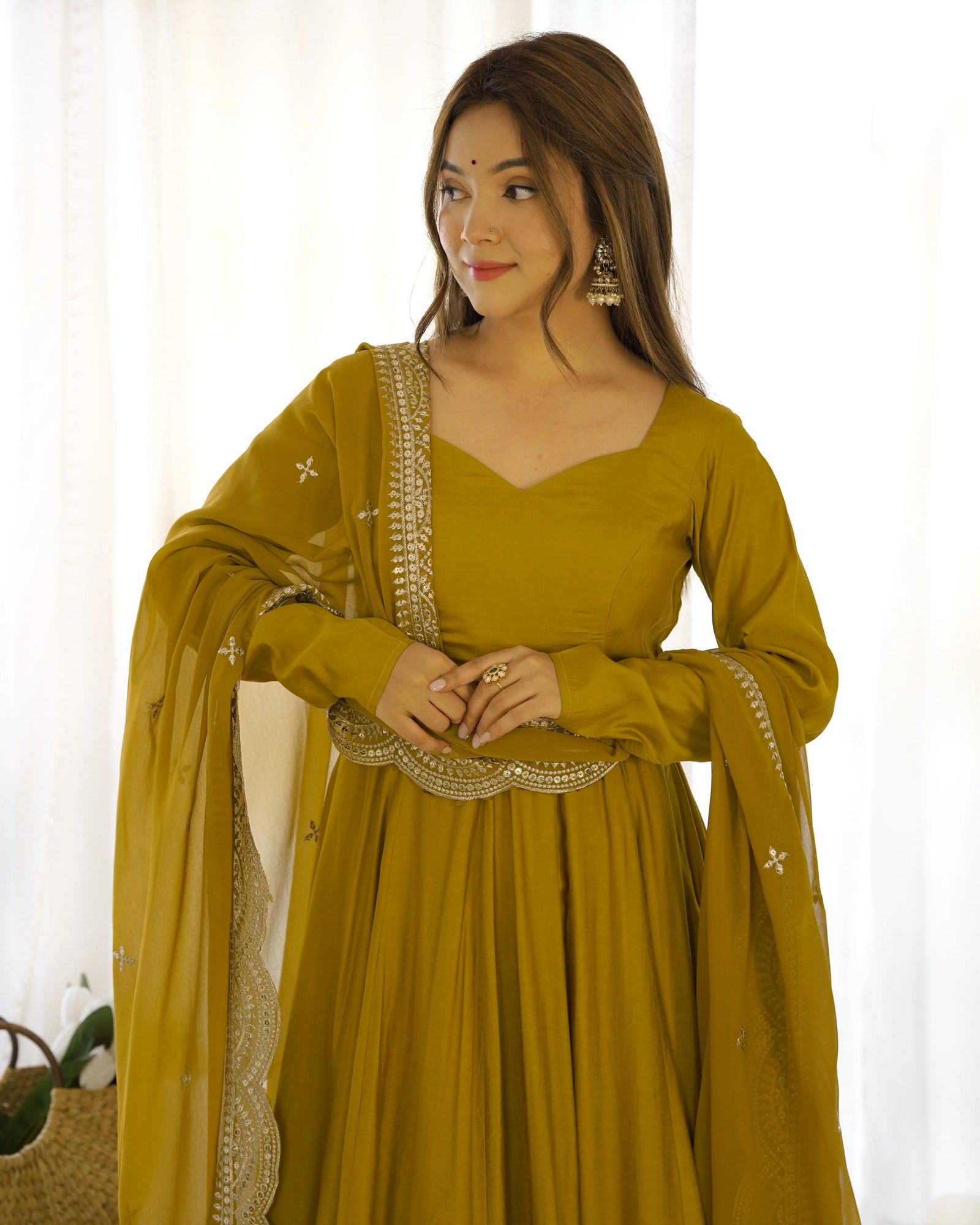 Woman wearing a mustard yellow traditional outfit with gold embroidery against a white curtain background