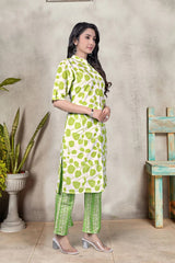 Woman wearing a green and white patterned outfit standing against a gray wall with a plant and wooden chair in the background.